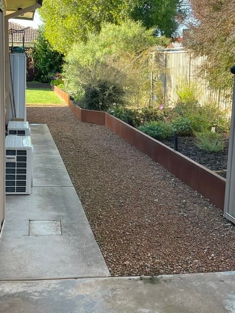 Corten steel retaining wall separating a rock walkway from a planted garden beside a house.
